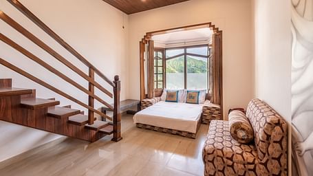 A spacious room near Bhagsunag Temple featuring a wooden staircase, a bed, a sofa, and a large window offering a mountain view – DLS Meghavan Resort.
